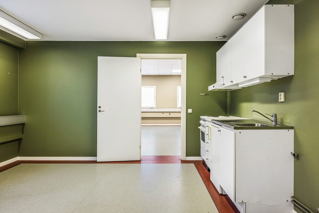 Ett litet kök med vita skåp, spis och diskho mot en grön vägg; en öppen dörr leder till ett starkt upplyst, tomt angränsande rum.