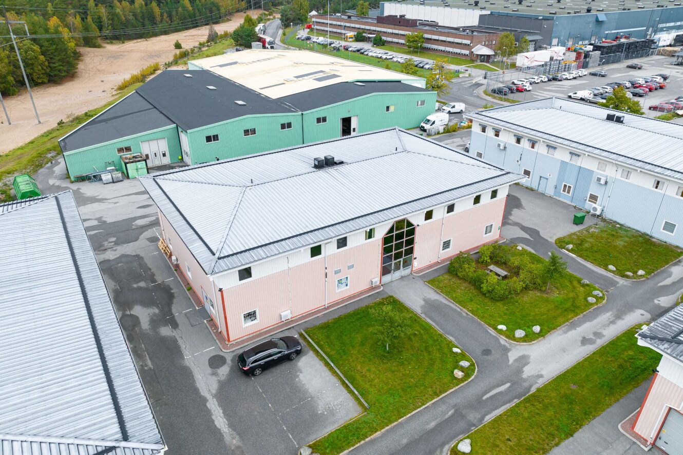 Flygfoto över ett industriområde med flera lagerbyggnader, en parkeringsplats och en svart bil parkerad utanför en ljusrosa byggnad.