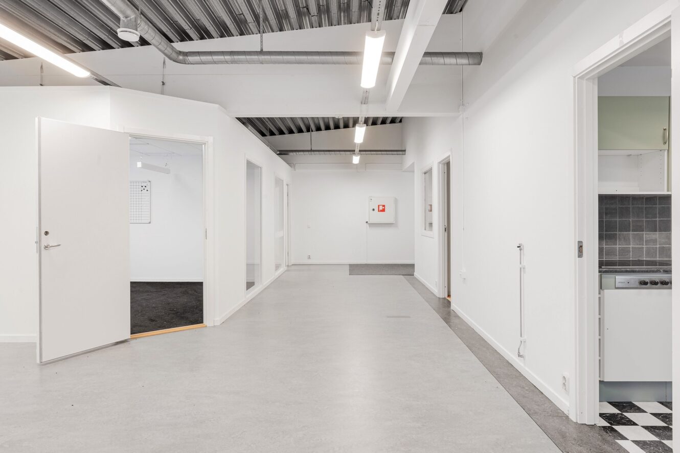 En ren, modern hall med vita väggar, öppna dörrar, takbelysning och synliga takkanaler; ett köksutrymme syns till höger.