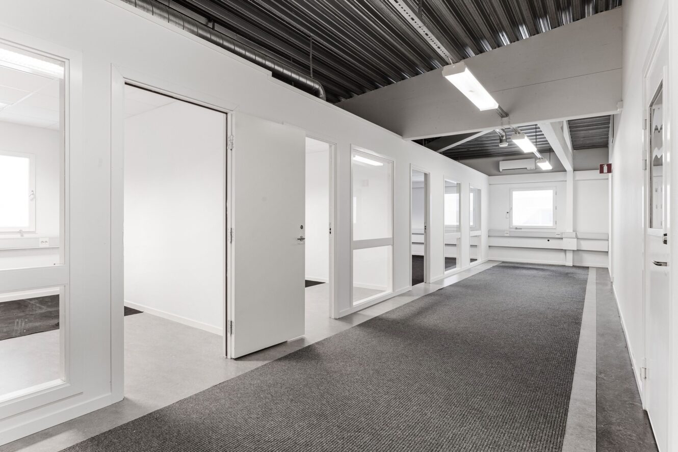En korridor med grå heltäckningsmatta och flera öppna vita dörrar som leder in till tomma kontorsrum med stora fönster och vita väggar.