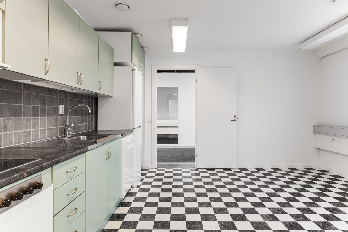Ett kök med mintgröna skåp, svarta bänkskivor, grå kakelplattor och ett svartvitrutigt golv. En dörr är öppen till ett angränsande rum.