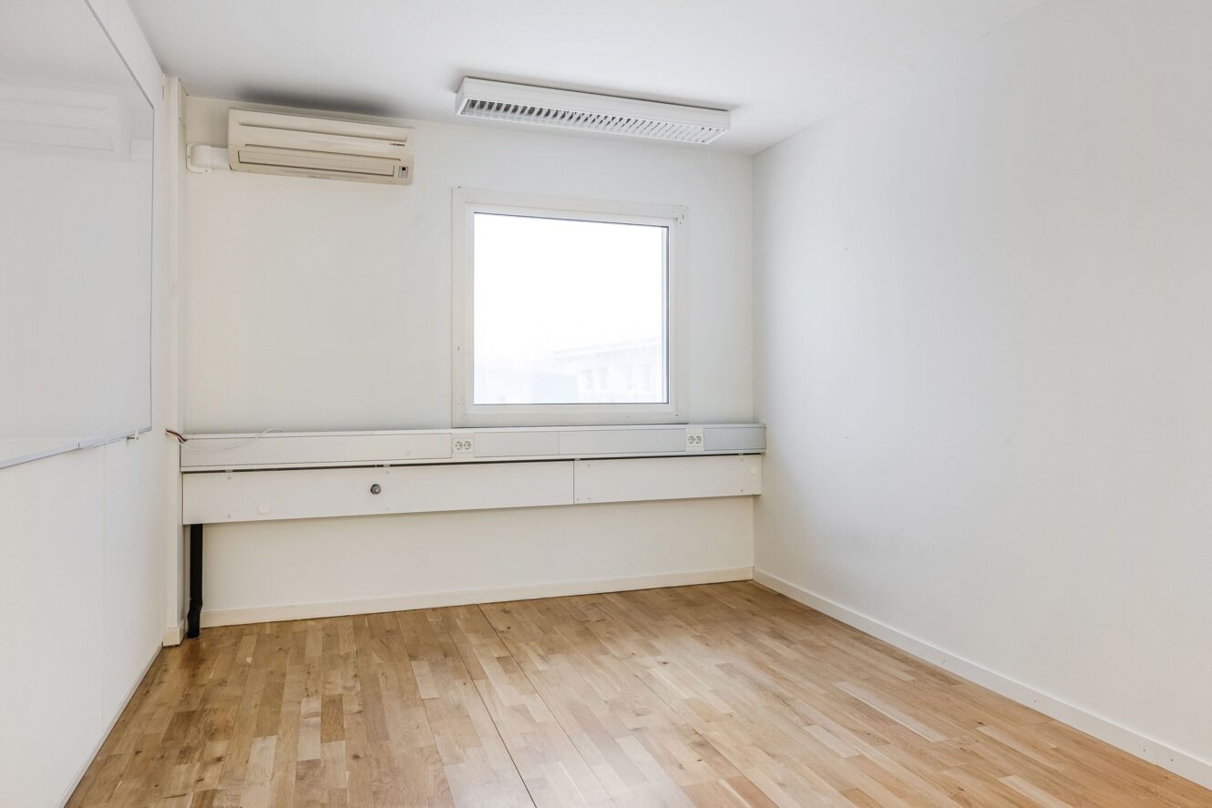 Ett tomt, omöblerat rum med vita väggar, ett enda fönster, trägolv och en väggmonterad luftkonditioneringsenhet.