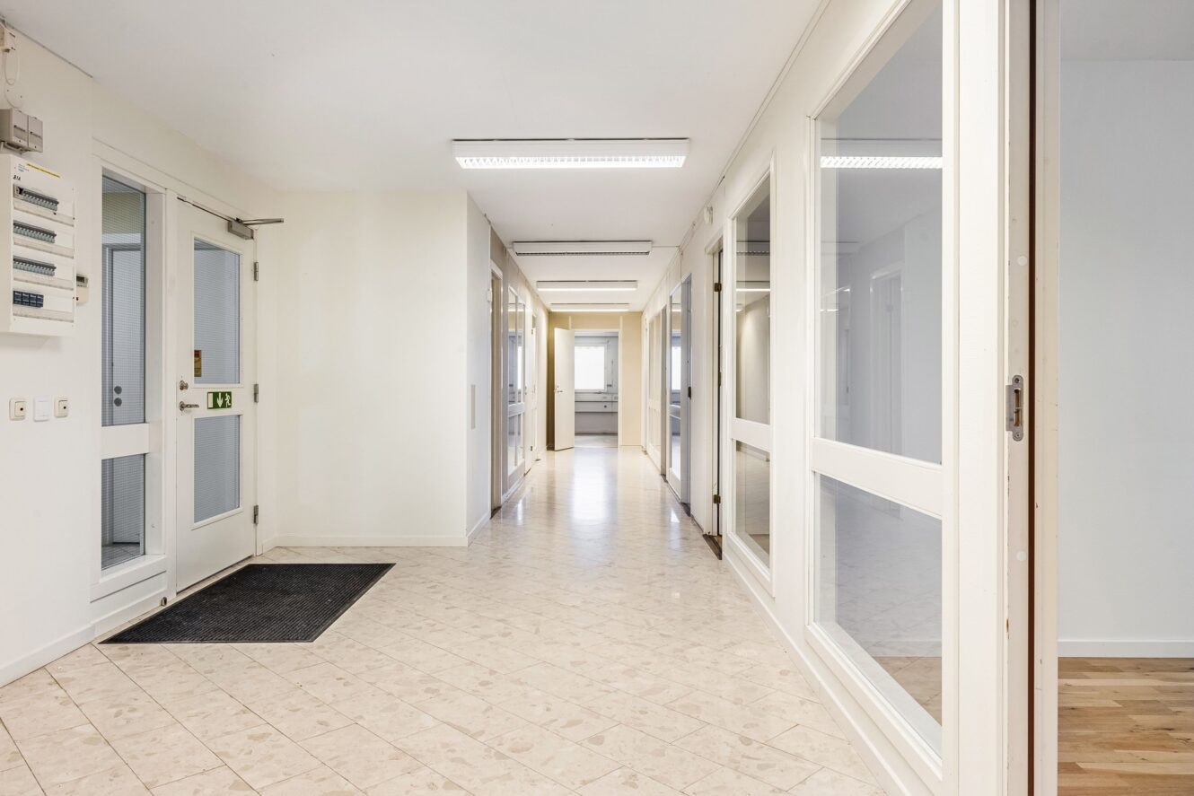 En ren, väl upplyst hall med klinkergolv, vita väggar, glasdörrar och fönster, som leder till ett rum med ett fönster längst bort.