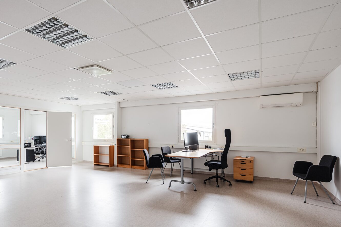 Ett rymligt, minimalistiskt kontor med ett stort skrivbord, en kontorsstol, två gäststolar, hyllor och fönster som släpper in naturligt ljus. Rummet är till största delen tomt och har vita väggar och takplattor.