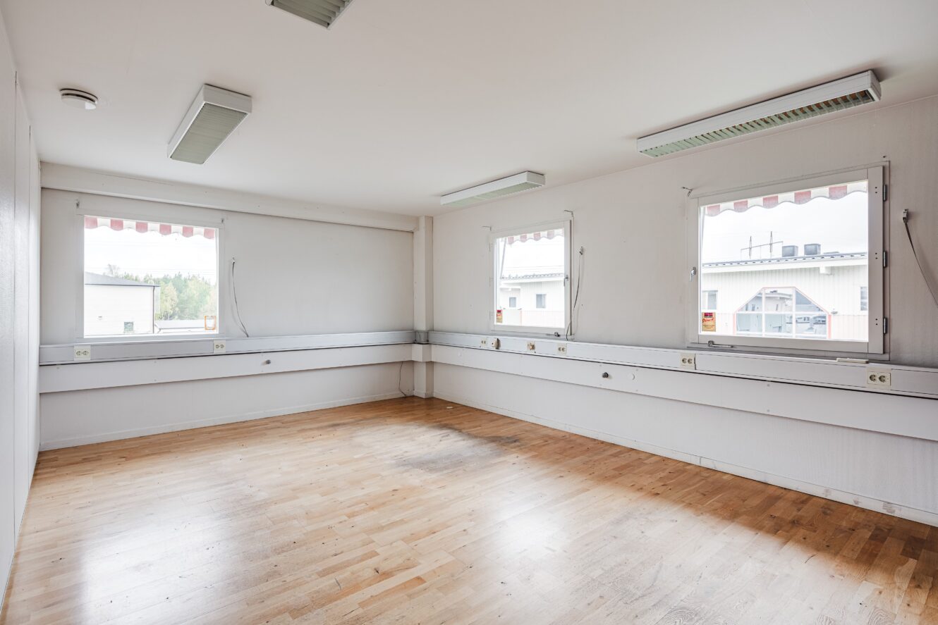 Tomt rum med ljusa väggar, trägolv, tre stora fönster, lysrör i taket och eluttag längs väggarna.