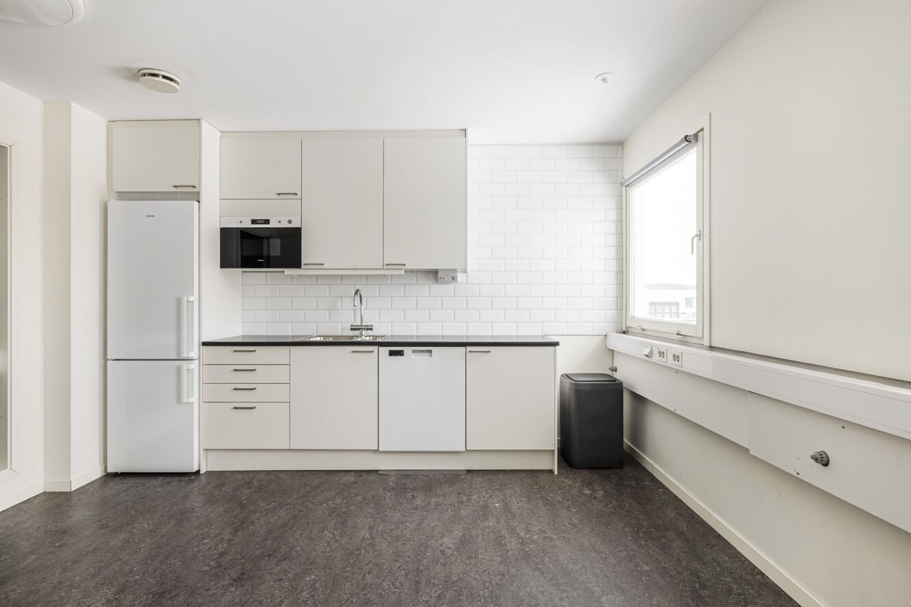 Ett modernt kök med vita skåp, kylskåp, mikrovågsugn, diskmaskin, diskho och ett fönster, allt mot en vit kakelplatta och grått golv.