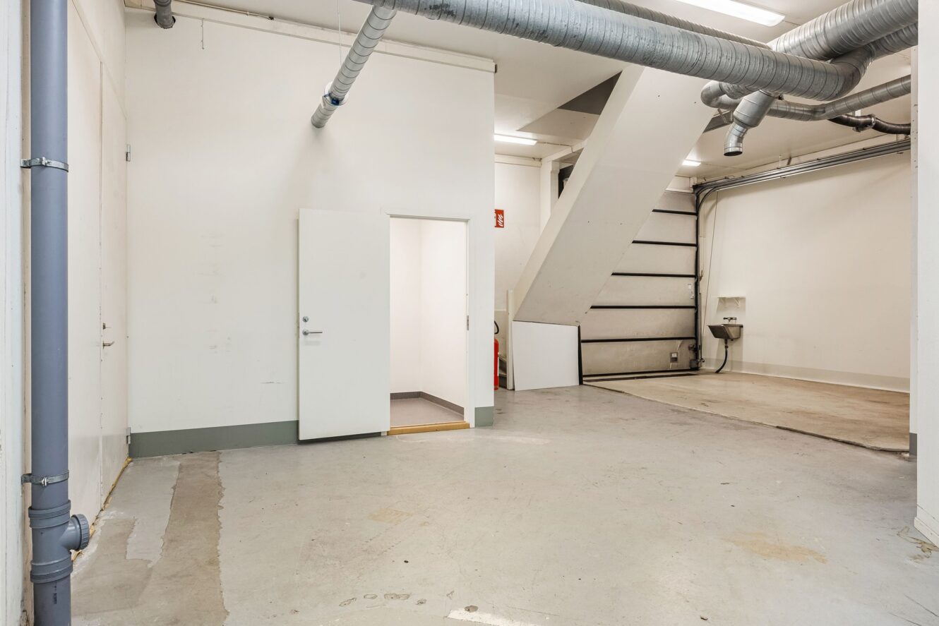 Industrilokal med synliga rör, öppen dörr till ett litet sidorum, en trappa, luftkanaler och en diskho på ett naket betonggolv.