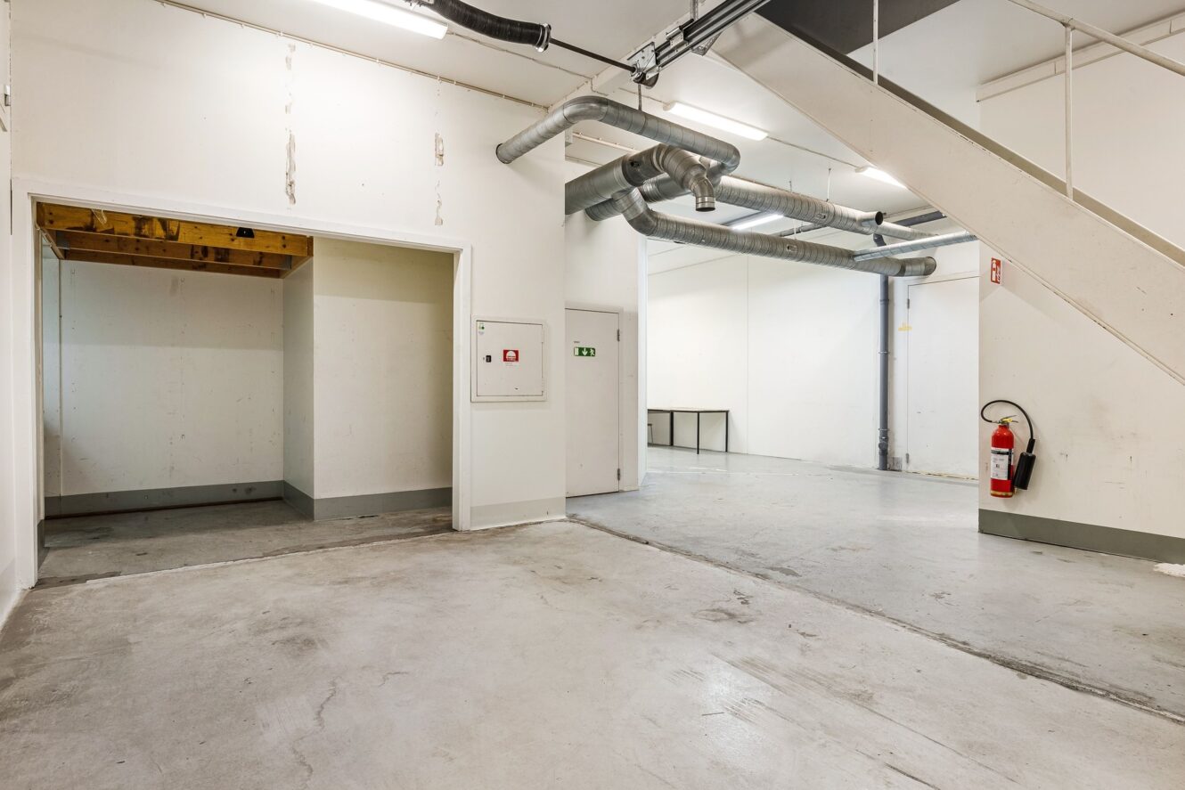 En tom industriinredning med vita väggar, synliga rörledningar, betonggolv, en trappa och en brandsläckare monterad på väggen.