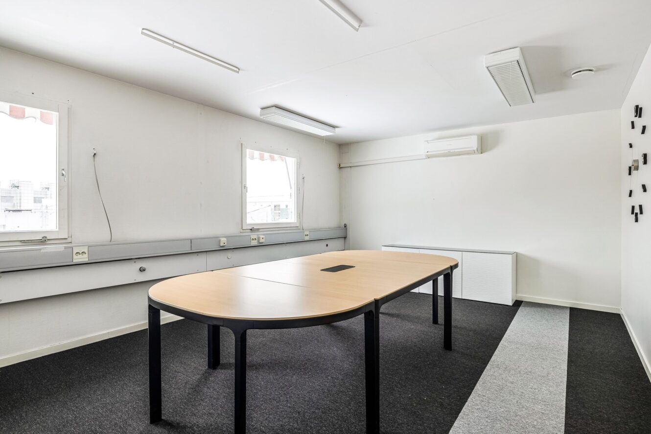 Ett vanligt konferensrum med två rektangulära bord ihopskjutna, svarta stolar, vita väggar och två fönster som släpper in naturligt ljus.