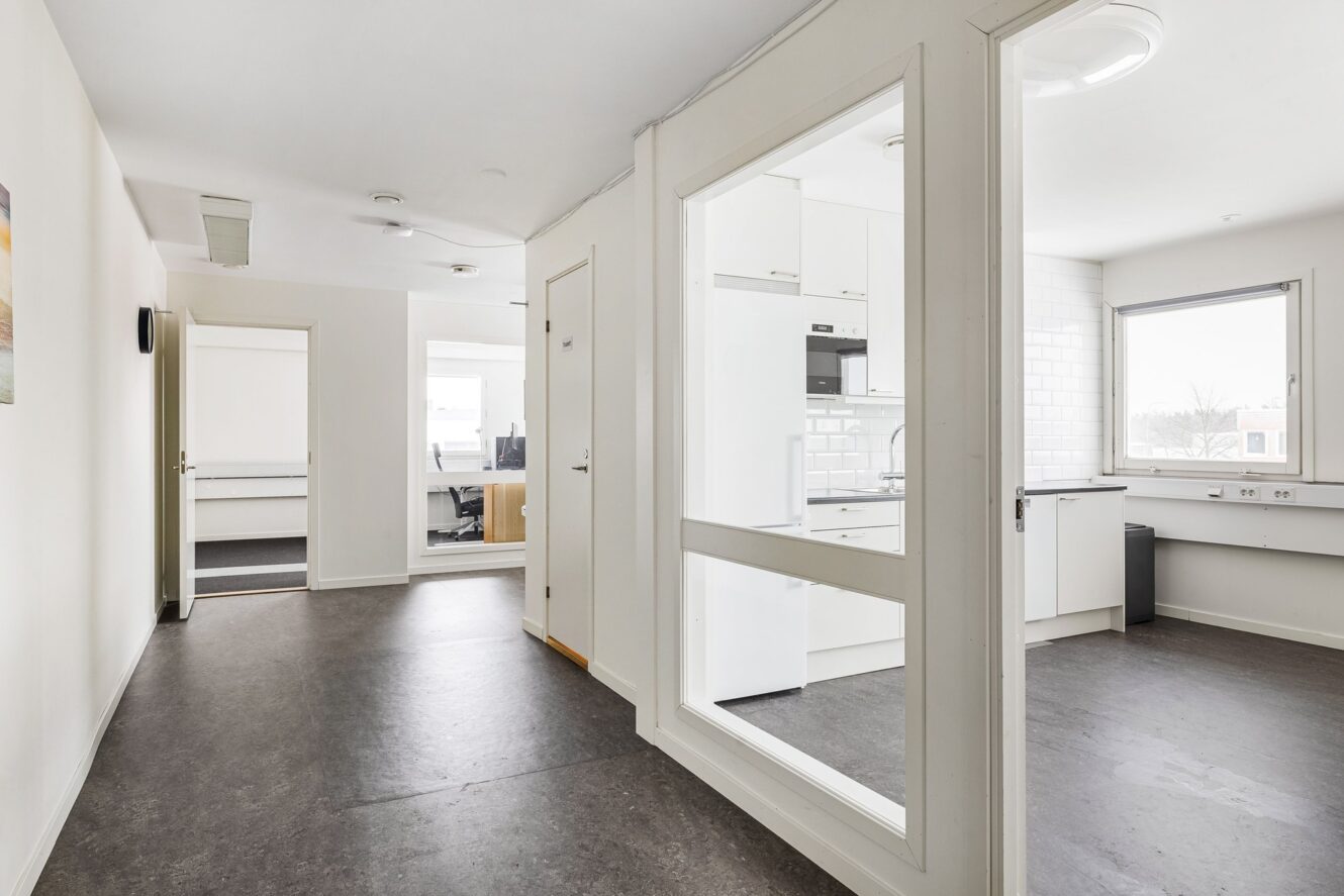 En ljus hall i en modern byggnad med dörrar som leder till ett kök, ett kontor och andra rum. Golven är mörka och väggarna är vita. Stora fönster släpper in naturligt ljus.