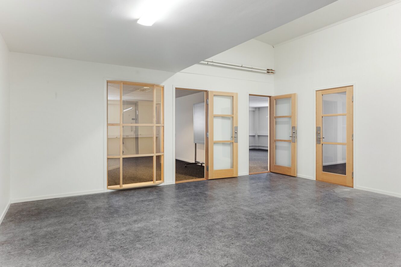 Ett tomt rum med grått golv, vita väggar, takbelysning och tre trädörrar med glaspaneler som leder till angränsande rum.