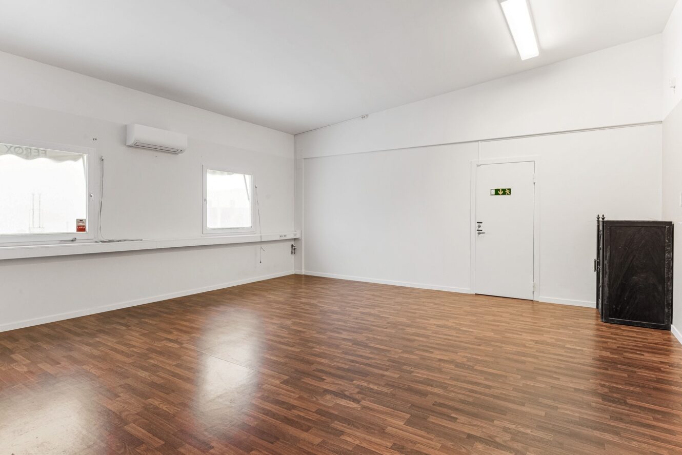 Ett tomt rum med vita väggar, trägolv, två fönster, en dörr med en utgångsskylt, lysrörsbelysning och ett svart skåp i hörnet.