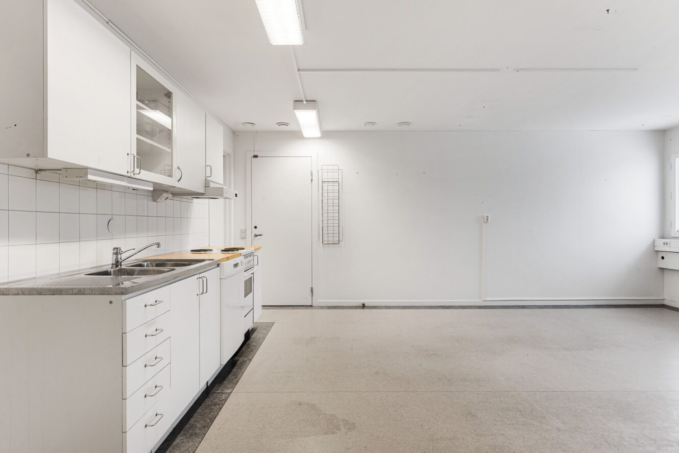 Ett minimalt, tomt kök med vita skåp, diskho, spis och kaklat stänkskydd mot vita väggar och golv.