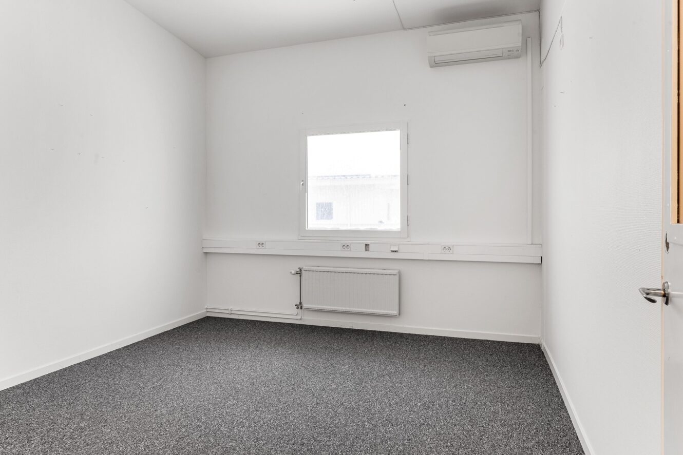 Tomt vitt rum med grå heltäckningsmatta, ett enda fönster, radiator, luftkonditioneringsenhet och eluttag på väggarna.