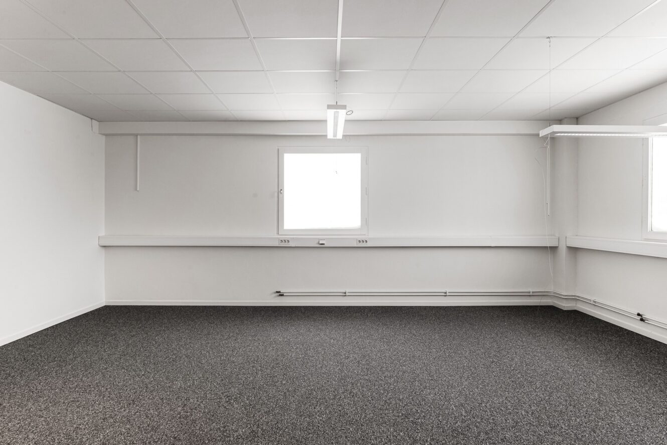 Tomt kontorsrum med vita väggar, grå matta, en taklampa och två fönster som släpper in naturligt ljus.