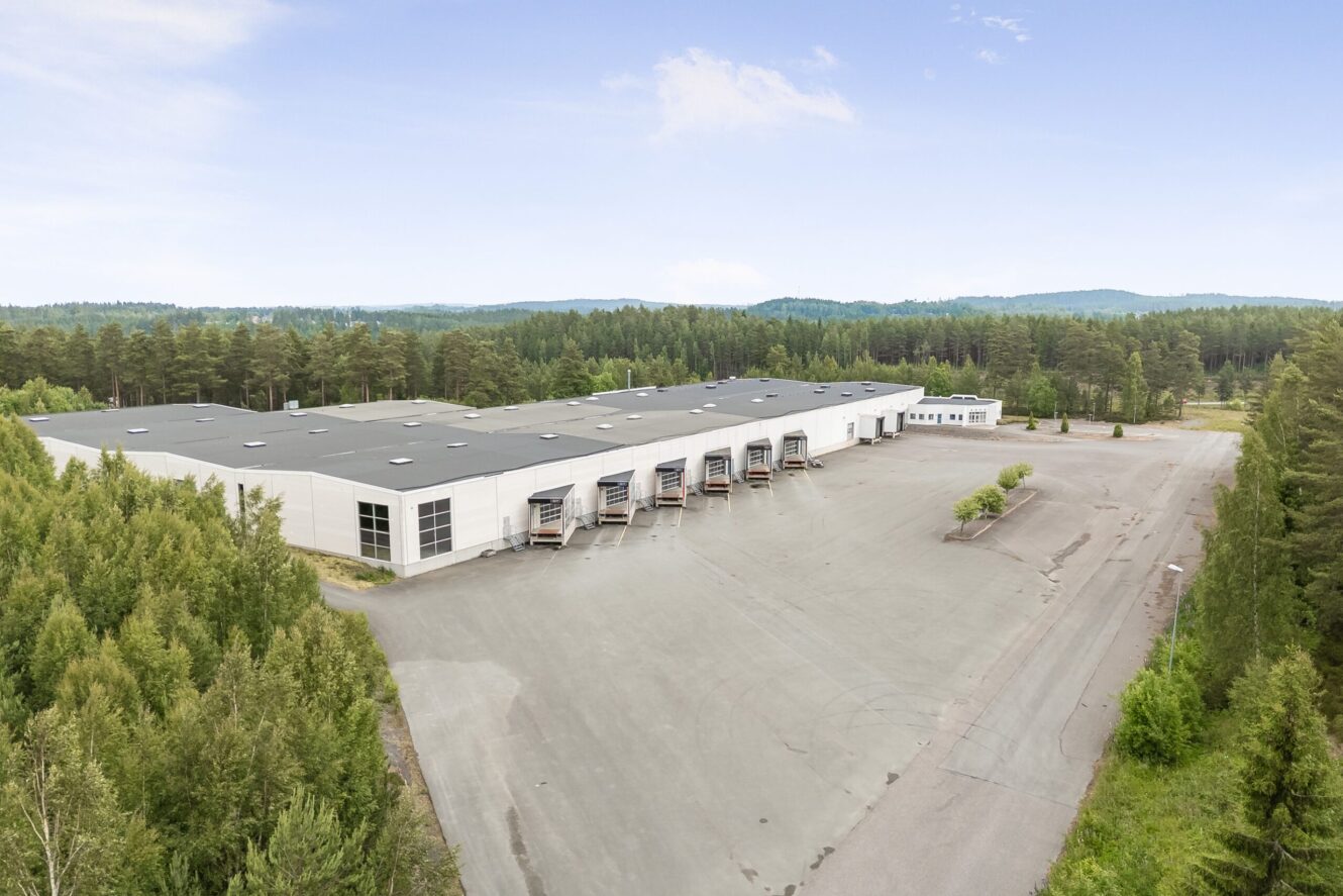 En stor lagerbyggnad med lastkajer, omgiven av träd och en stor tom parkeringsplats under en klar himmel.