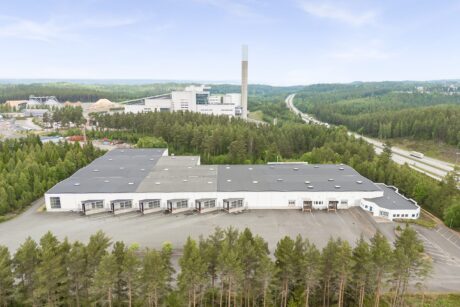 Flygfoto över ett stort industrilager med flera lastkajer, omgivet av träd, vägar och ytterligare industribyggnader i bakgrunden.