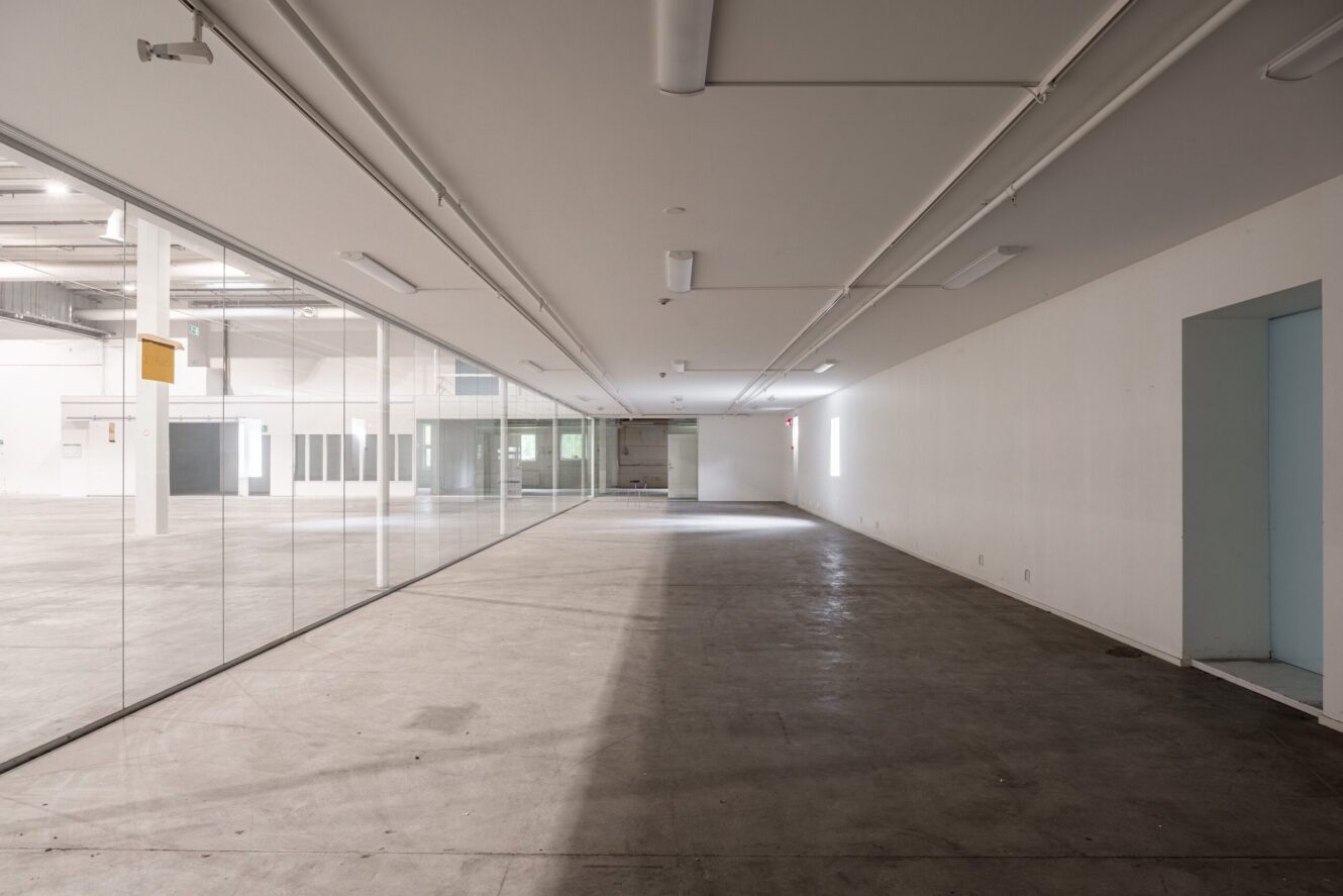 Tomt industriområde med betonggolv, vita väggar och taklampor; glasvägg till vänster och dörröppningar till höger; solljus kommer in från bortre änden.