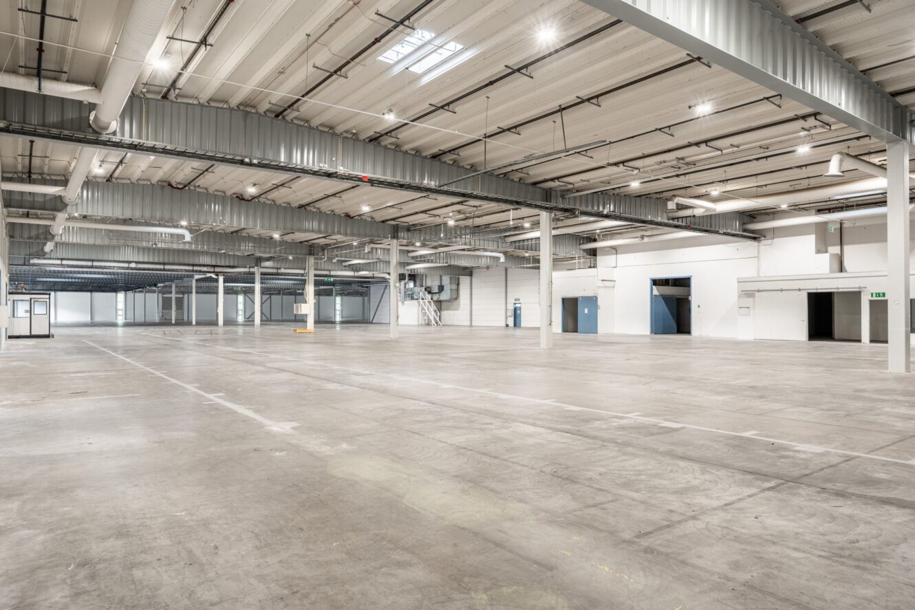 Stort tomt industrilager med betonggolv, synliga takbjälkar, ventilationskanaler och flera entrédörrar längs väggarna.