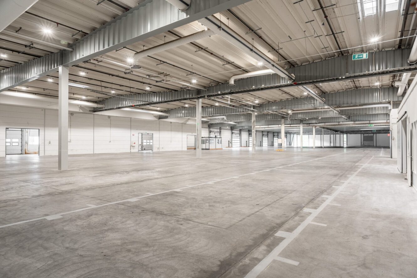 Rymlig, tom industrilagerlokal med betonggolv, högt i tak, synliga kanaler och stora fönster och dörrar längs väggarna.