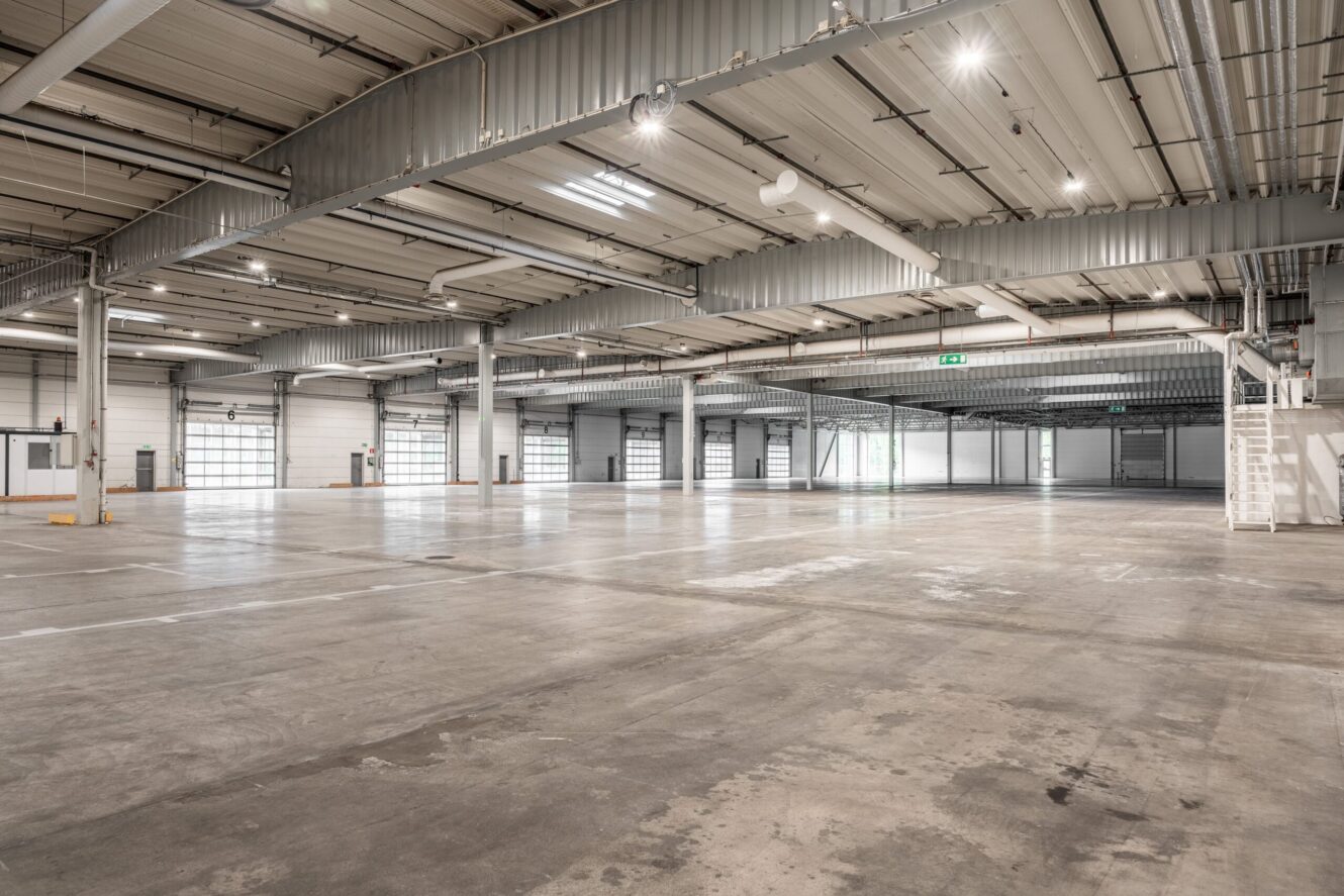 Stort, tomt industrilager med betonggolv, högt i tak, synliga bjälkar och flera takskjutportar längs väggarna.