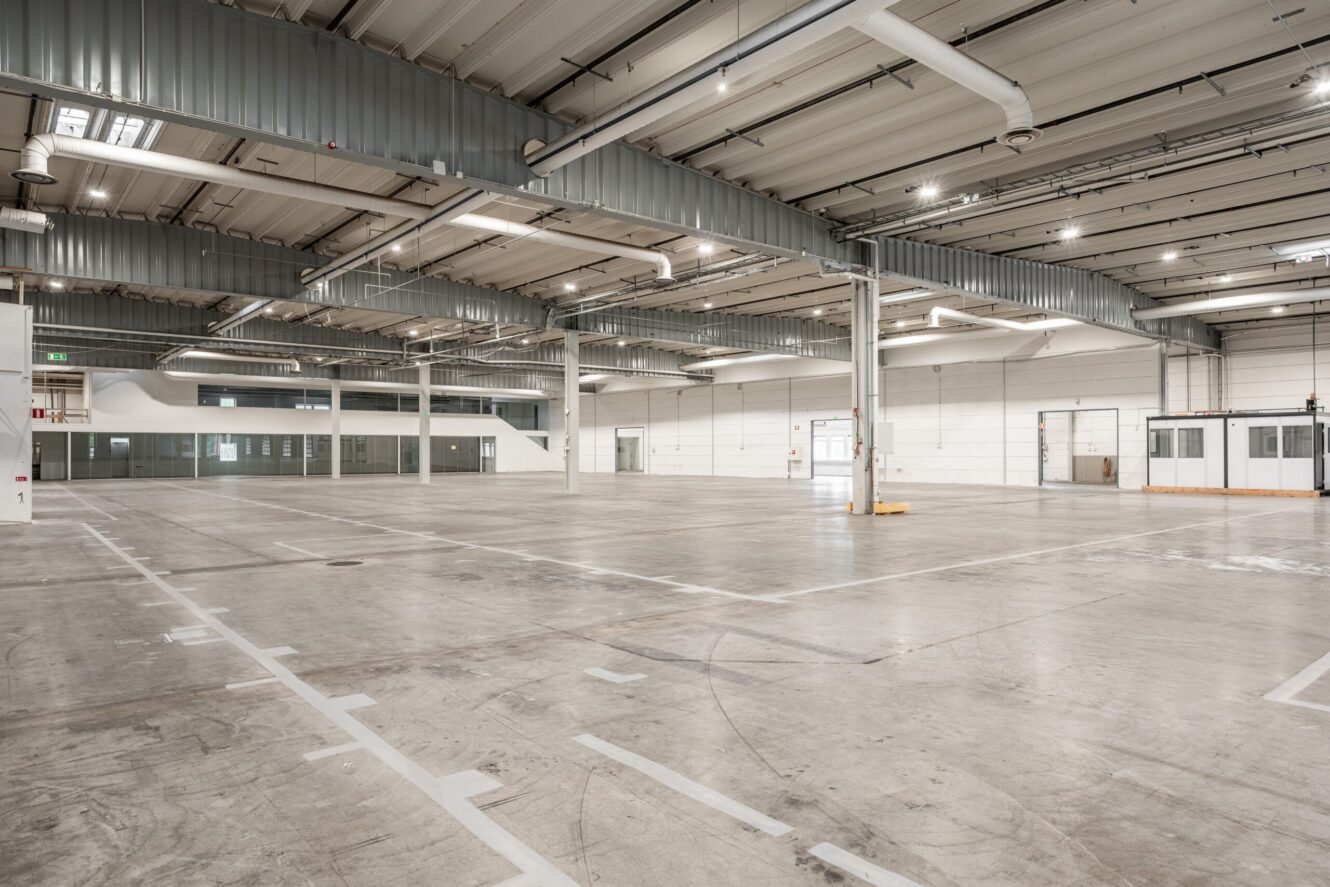 Stort, tomt industrilager med betonggolv, högt i tak, synliga ventilationskanaler och flera slutna kontorsutrymmen längs väggarna.