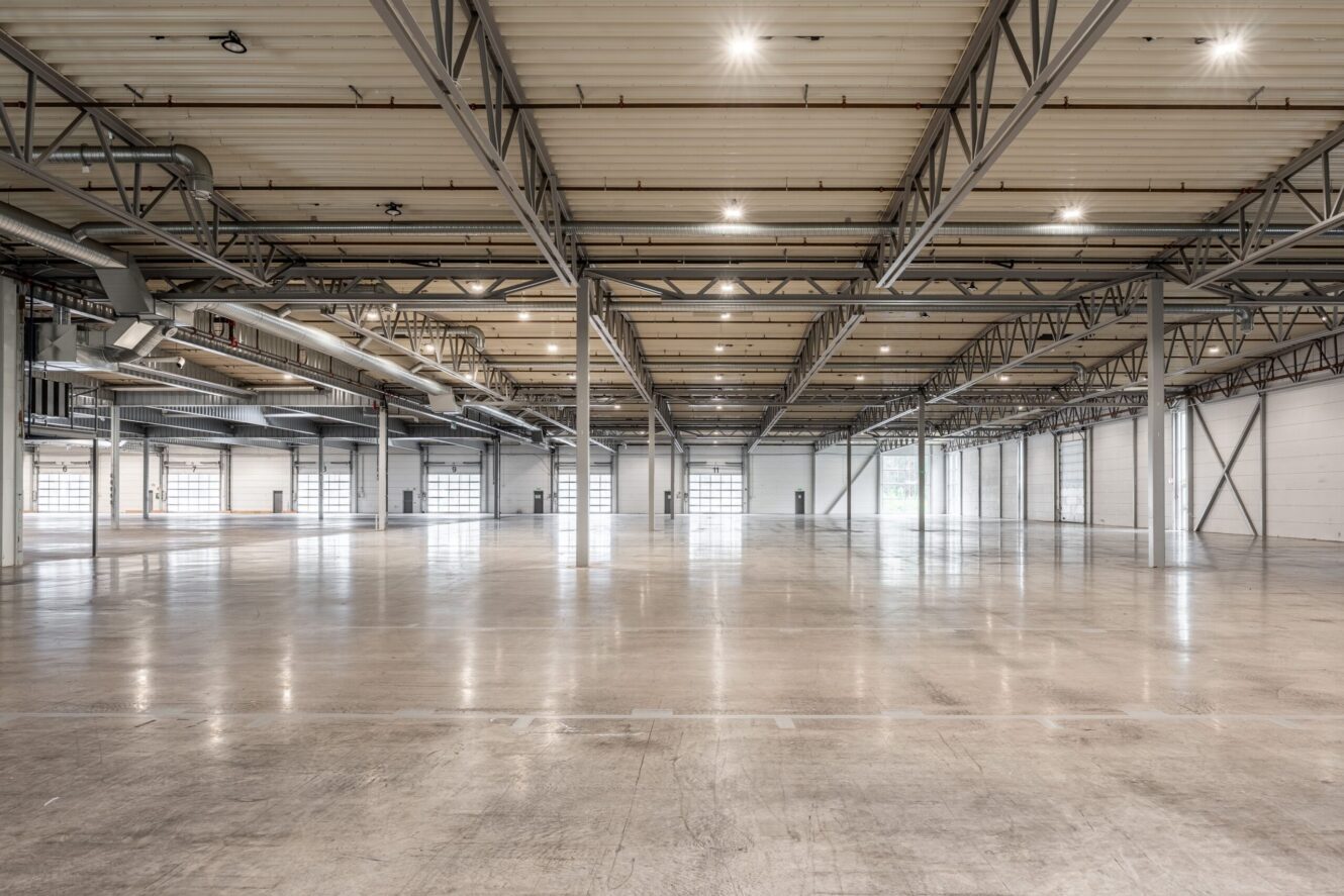 Stort, tomt industrilager med betonggolv, synliga stålbalkar och takbelysning; flera stora garageportar längs väggarna.