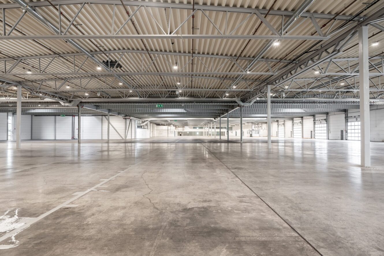 Stort, tomt industrilager med betonggolv, högt i tak, metallbalkar och flera lastkajsdörrar längs väggarna.