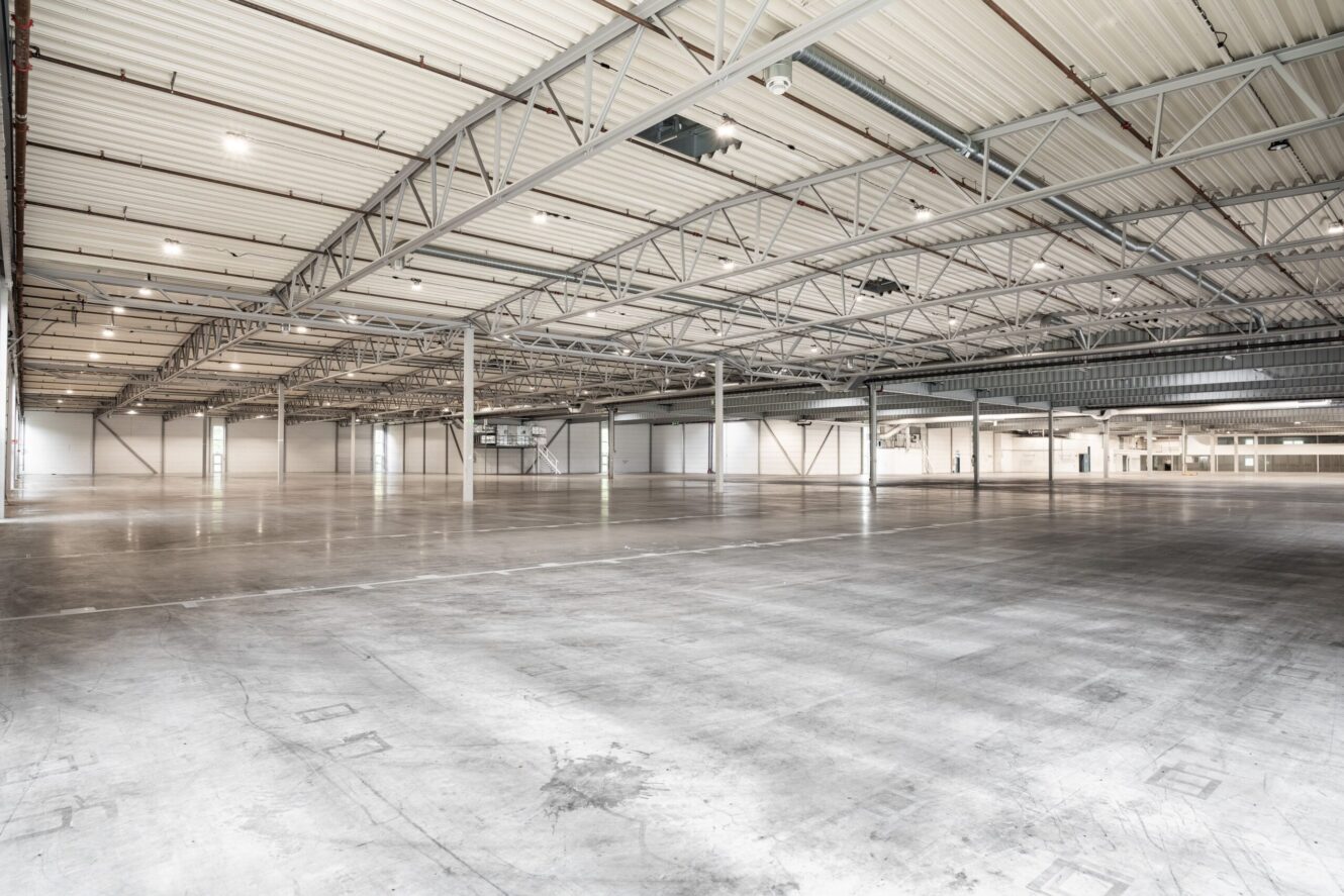 Stort, tomt industrilager med högt i tak, synliga bjälkar och betonggolv, upplyst av naturligt och artificiellt ljus.