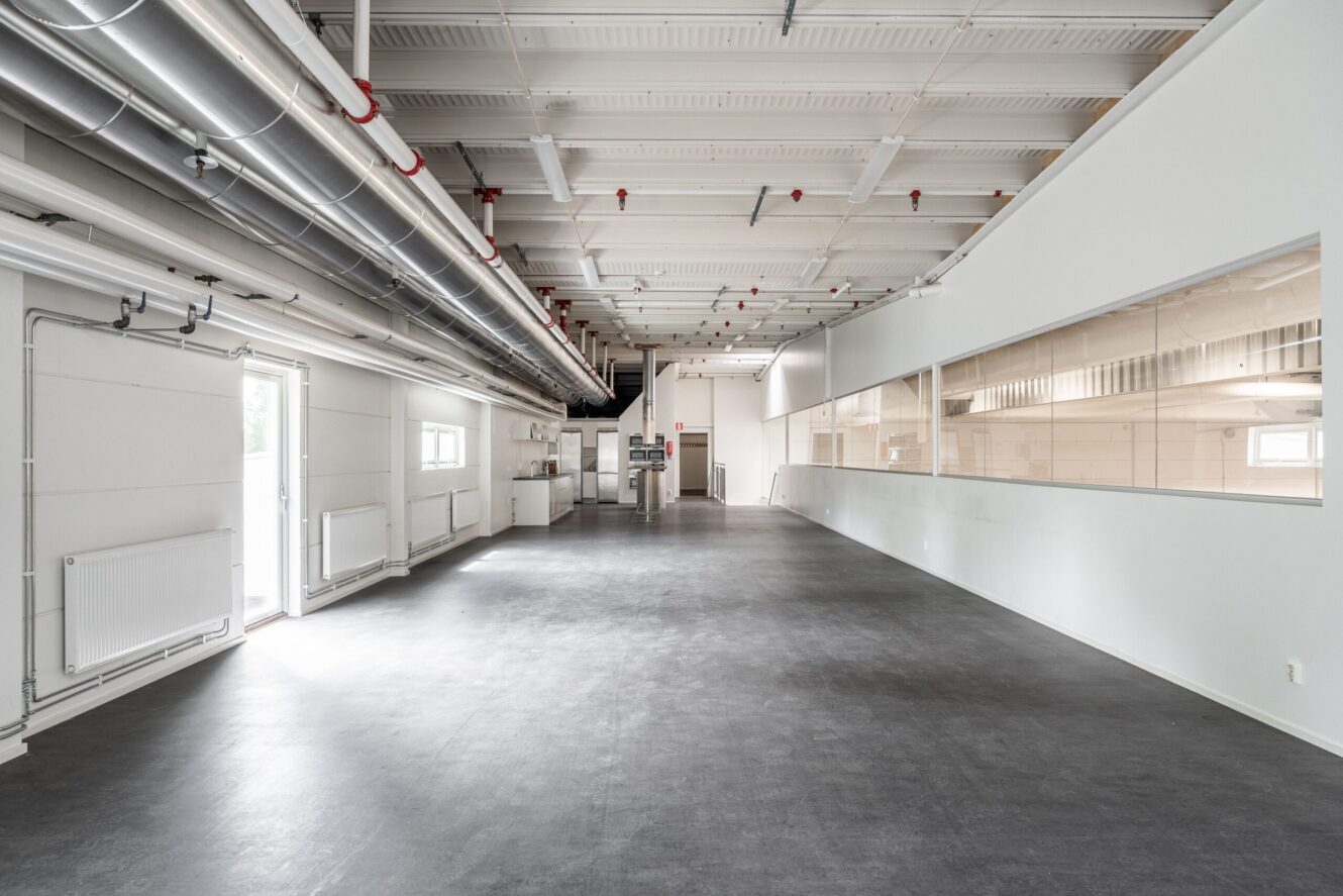 Tom industriinredning med vita väggar, stora fönster, synliga rör i taket och grått golv. Till vänster finns en dörr som är öppen utåt.