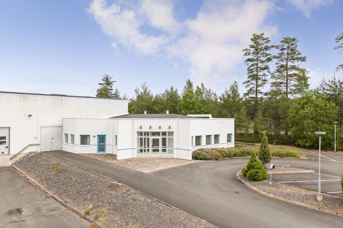 En modern vit kommersiell byggnad med blå accenter, stor glasentré, omgivande trottoar och närliggande träd under en delvis molnig himmel.