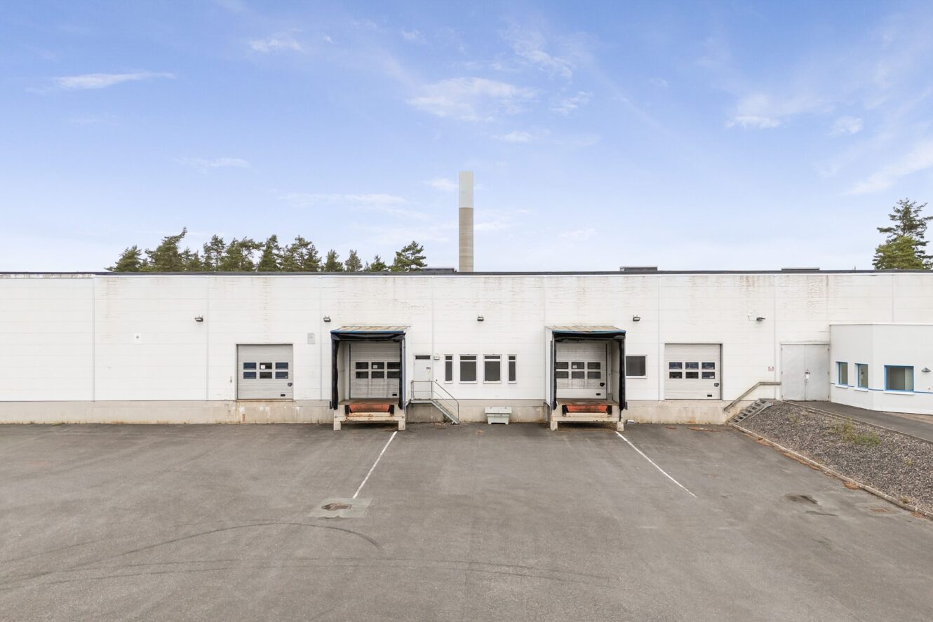 En lagerlokal med två lastkajer, en garageport och en tom asfalterad tomt under en klar himmel.