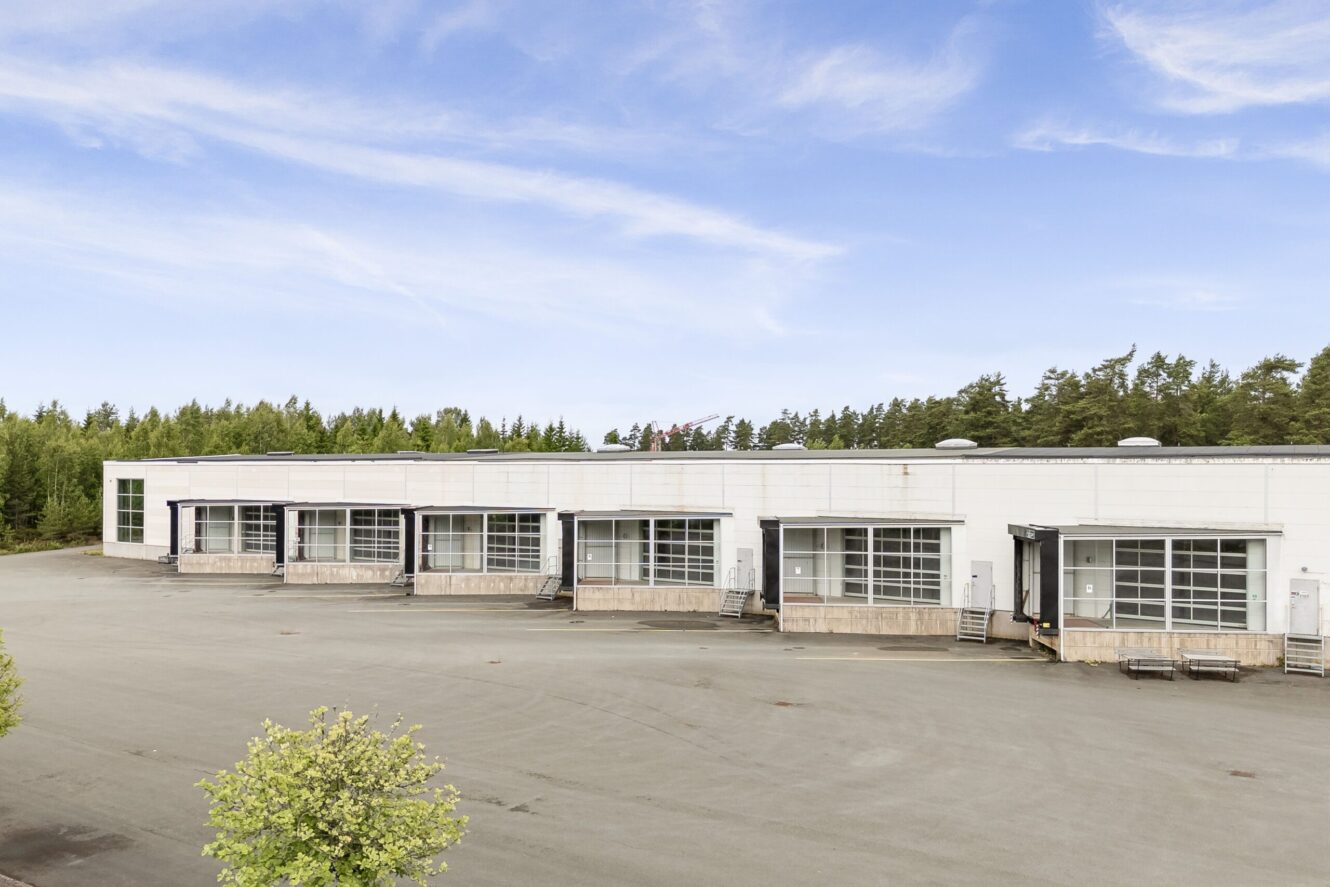 Ett stort, vitt industrilager med flera lastbryggor på en tom, asfalterad tomt med träd i bakgrunden under en delvis molnig himmel.