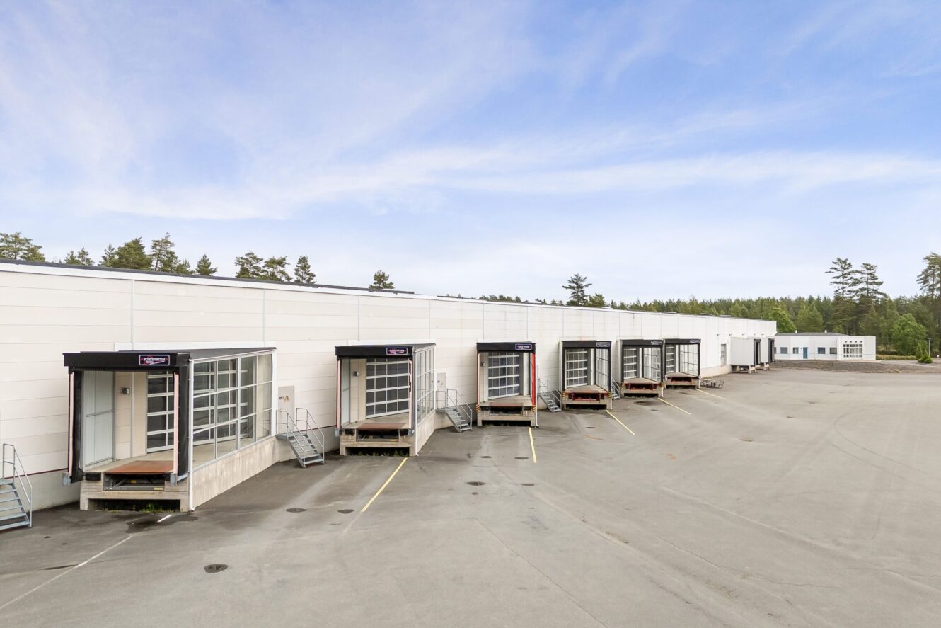 En rad lastbryggor vid ett stort vitt industriellt lager, med en tom asfalterad tomt framför och träd i bakgrunden.