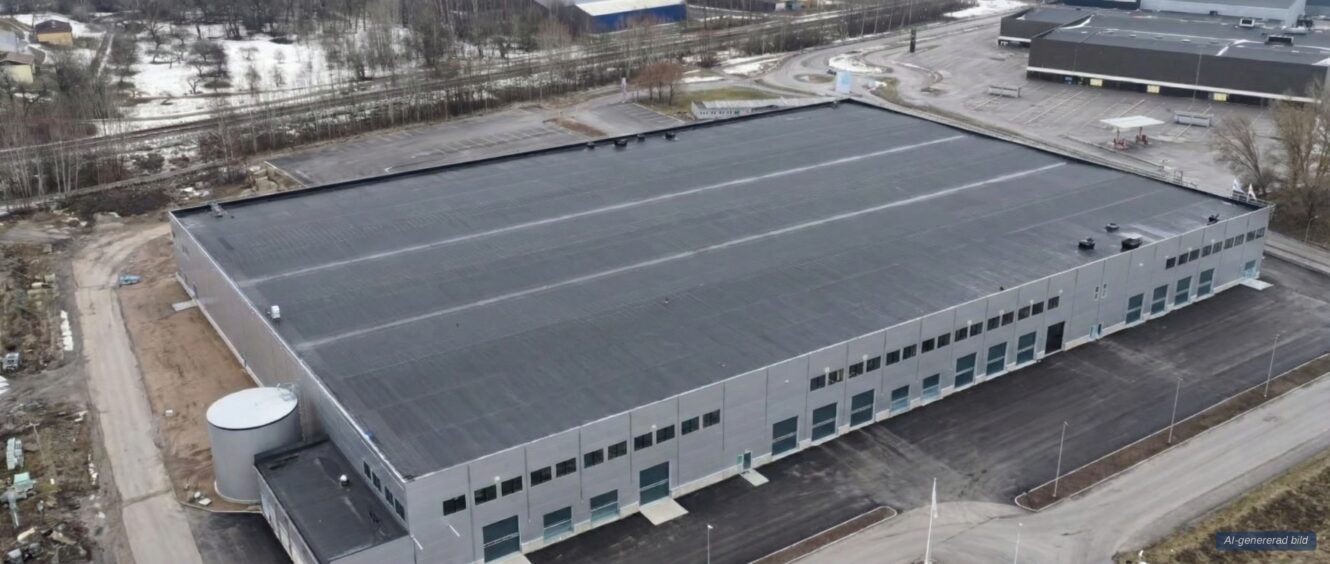 En stor, rektangulär industribyggnad med ett platt svart tak ligger på en i stort sett tom parkeringsplats, omgiven av vägar och gles vegetation.