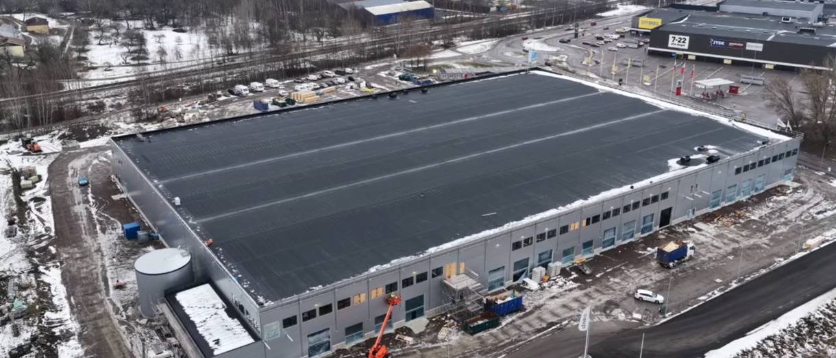 Flygfoto över en stor rektangulär industribyggnad med platt svart tak, omgiven av snö, fordon och närliggande kommersiella byggnader.