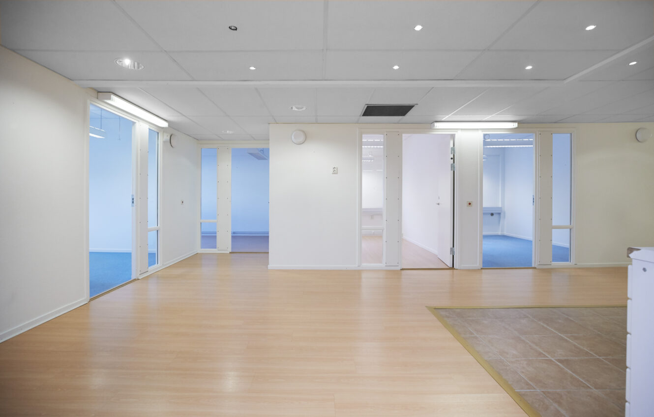 Tom modern kontorsinredning med ljust trägolv, vita väggar, infälld belysning, glasdörrar och en kaklad sektion till höger.