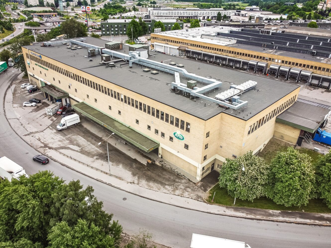 Ett stort industrilager med lastbryggor och parkerade lastbilar, omgivet av träd och andra industribyggnader.