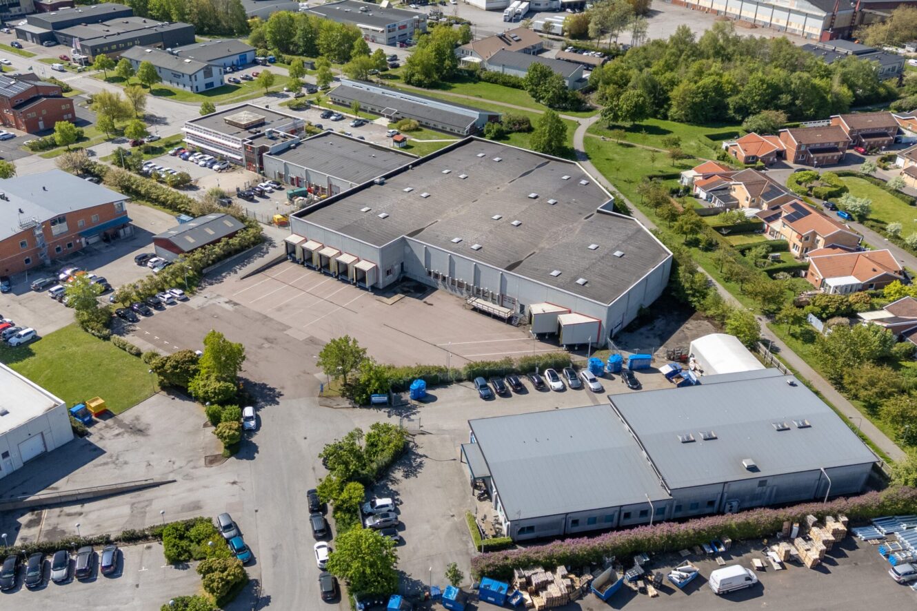 Flygfoto över ett industrikomplex med lagerlokaler, lastkajer, parkerade bilar och närliggande bostadshus omgivna av grönska.