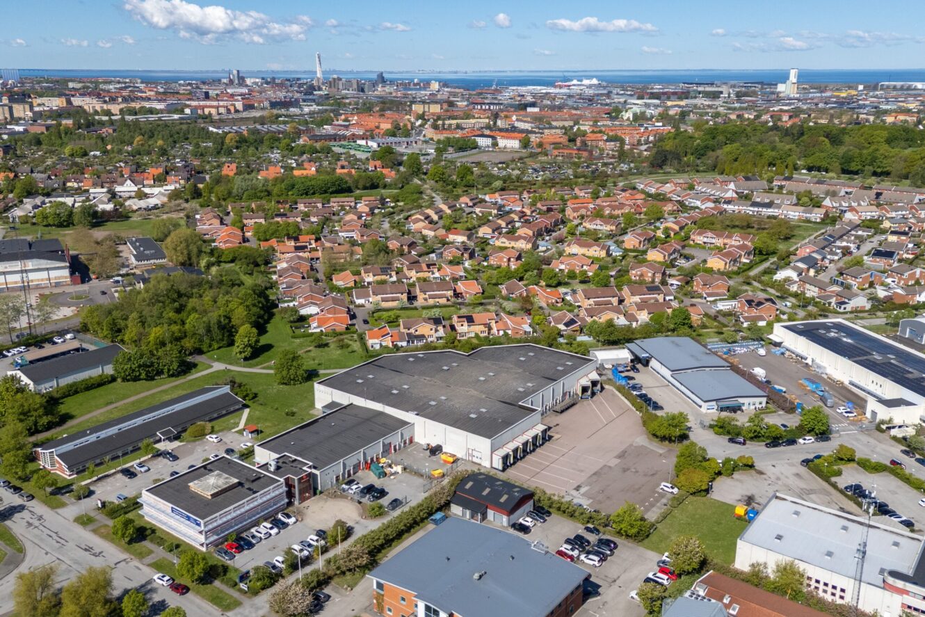 Flygfoto över ett förortsområde med rader av hus, kommersiella byggnader, parkeringsplatser och en stadsbild som syns i bakgrunden.