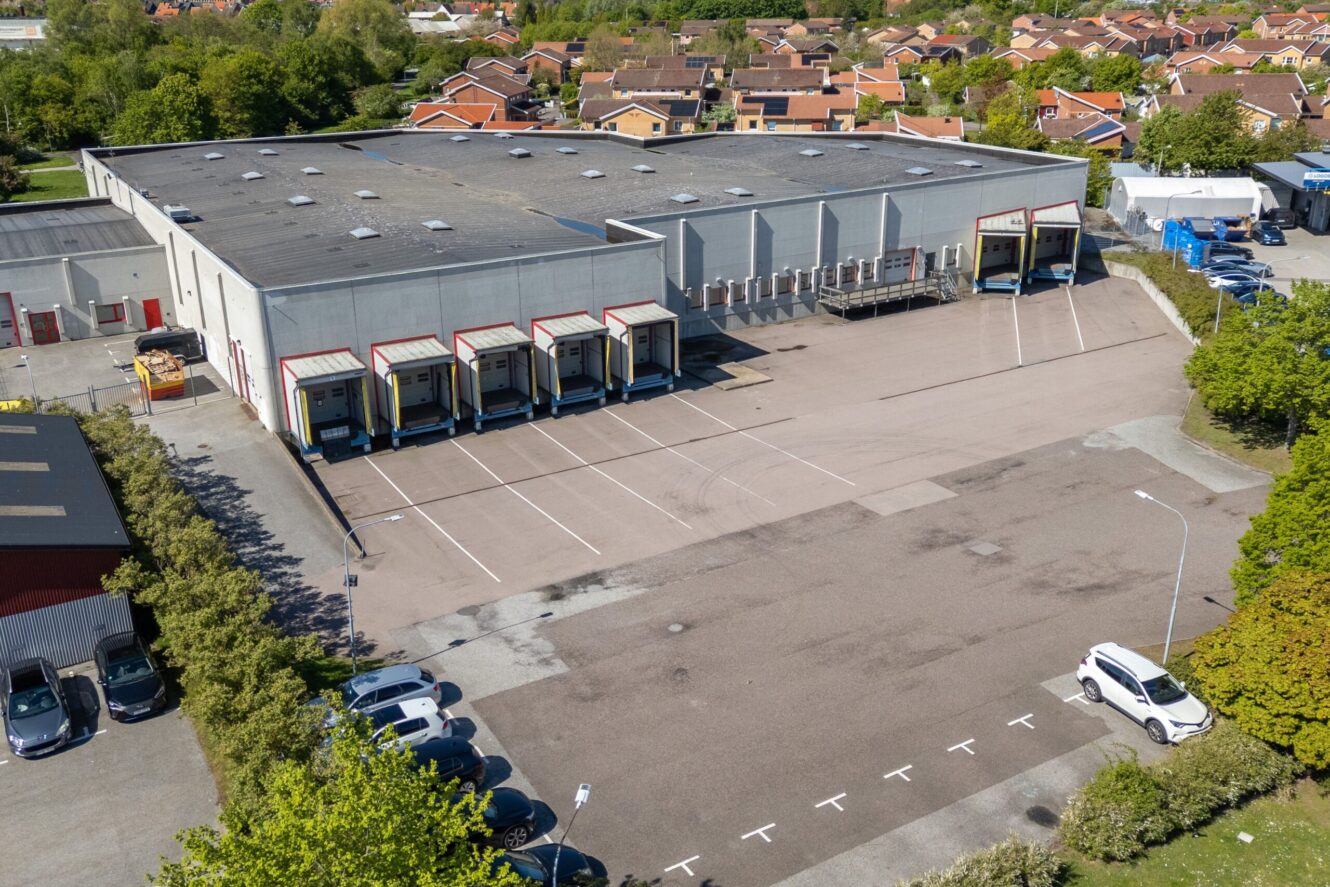 Flygfoto över en stor lagerlokal med flera lastkajer, flera parkerade bilar och ett bostadsområde i bakgrunden.