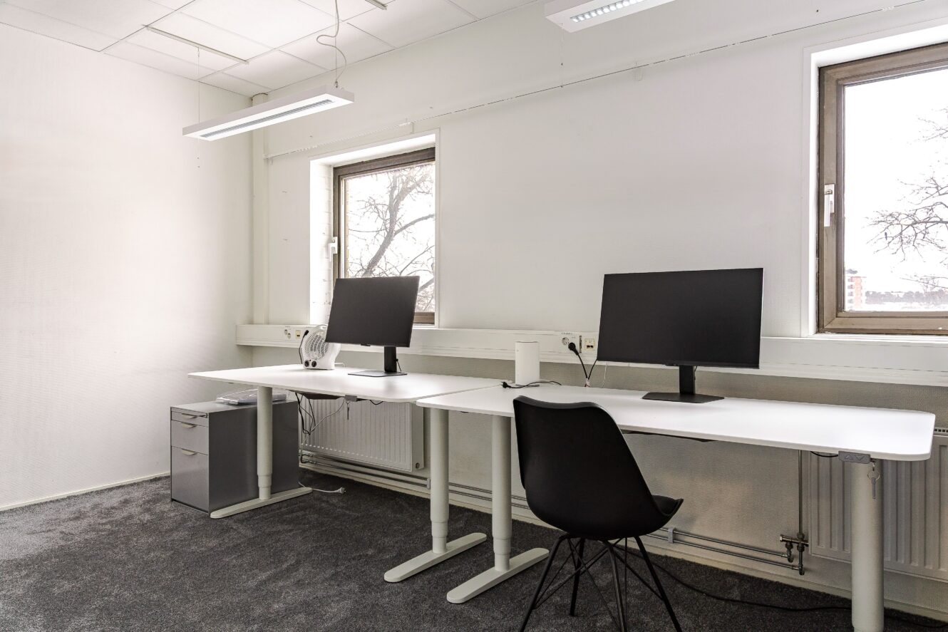 Ett minimalistiskt kontor med två skrivbord, två bildskärmar, en enda svart stol och två fönster som släpper in naturligt ljus.