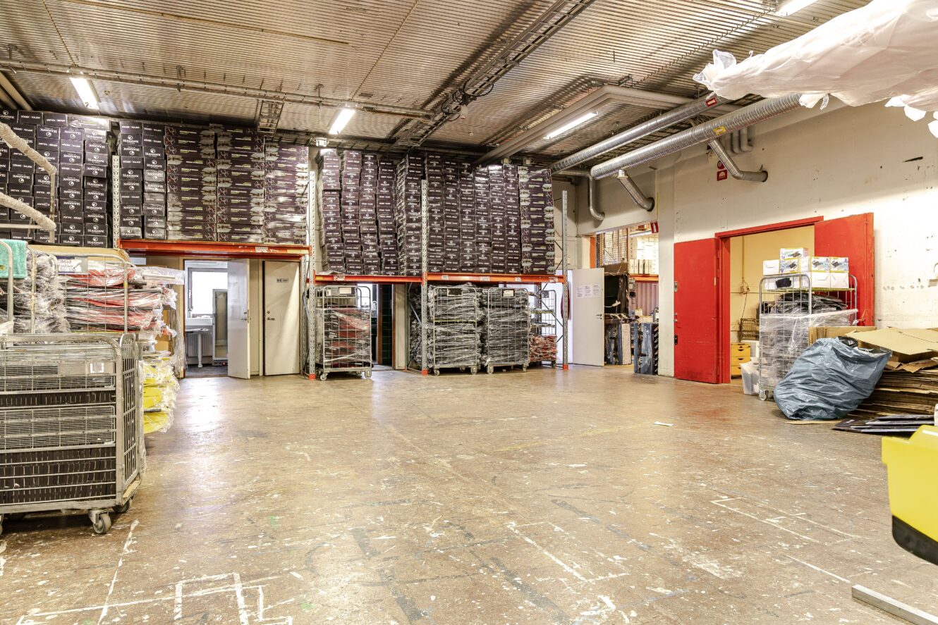 Lagerinredning med staplade lådor på höga hyllor, vagnar med varor, utspridda förpackningsmaterial och metalltak med synliga rör.