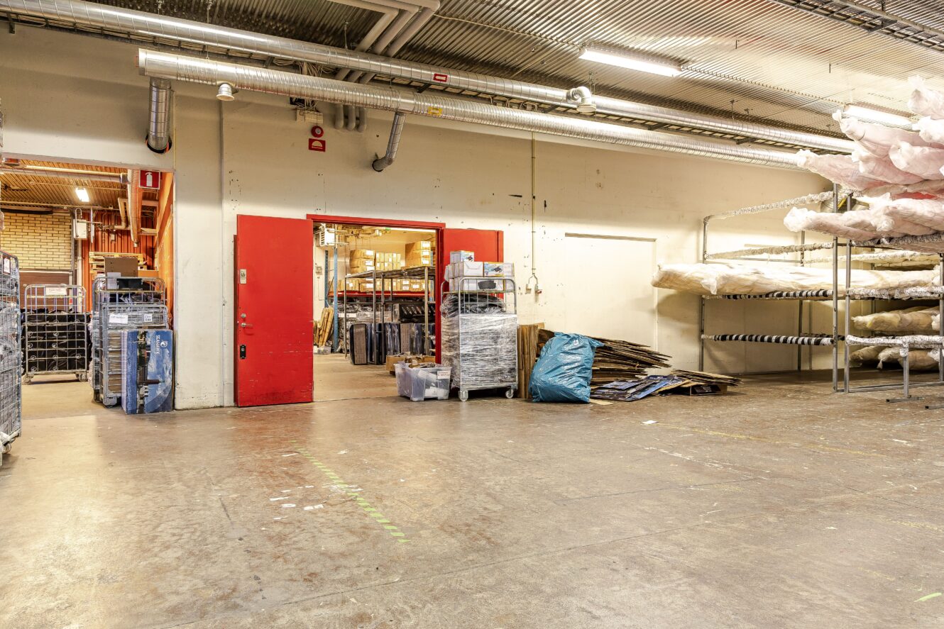 Lagerlokal med röda dubbeldörrar, metallhyllor med stora materialrullar och vagnar staplade mot väggen på ett betonggolv.