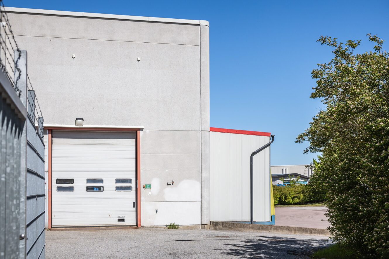 En ljusgrå industribyggnad med en stängd garageport ligger intill ett metallstaket och ett grönt träd under en klarblå himmel.