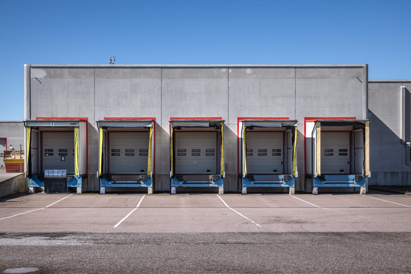 Fem tomma lastkajer med stängda takskjutportar vid ett lager, under en klarblå himmel.