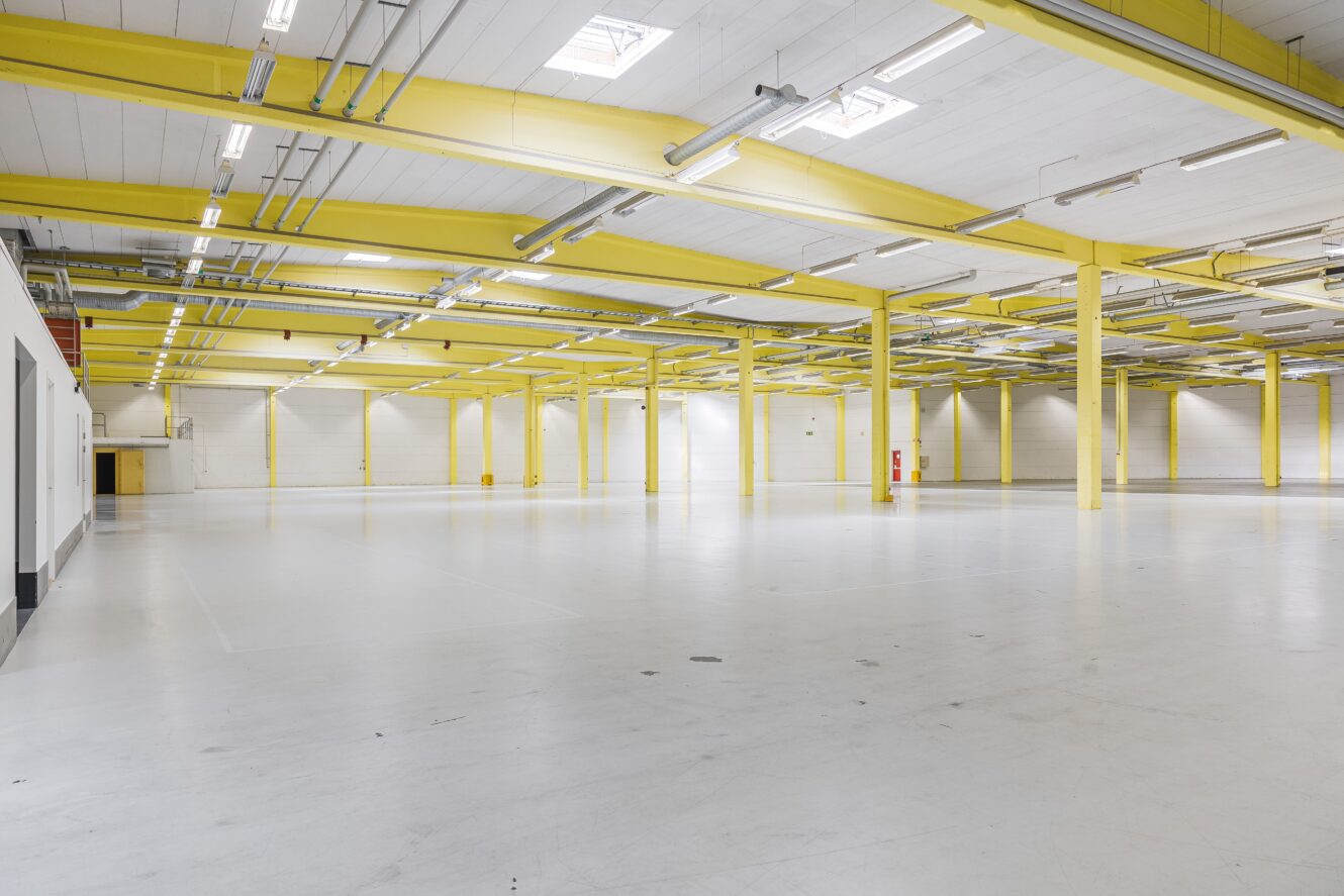 Rymlig, tom industrihall med gula balkar, vita golv och taklampor.