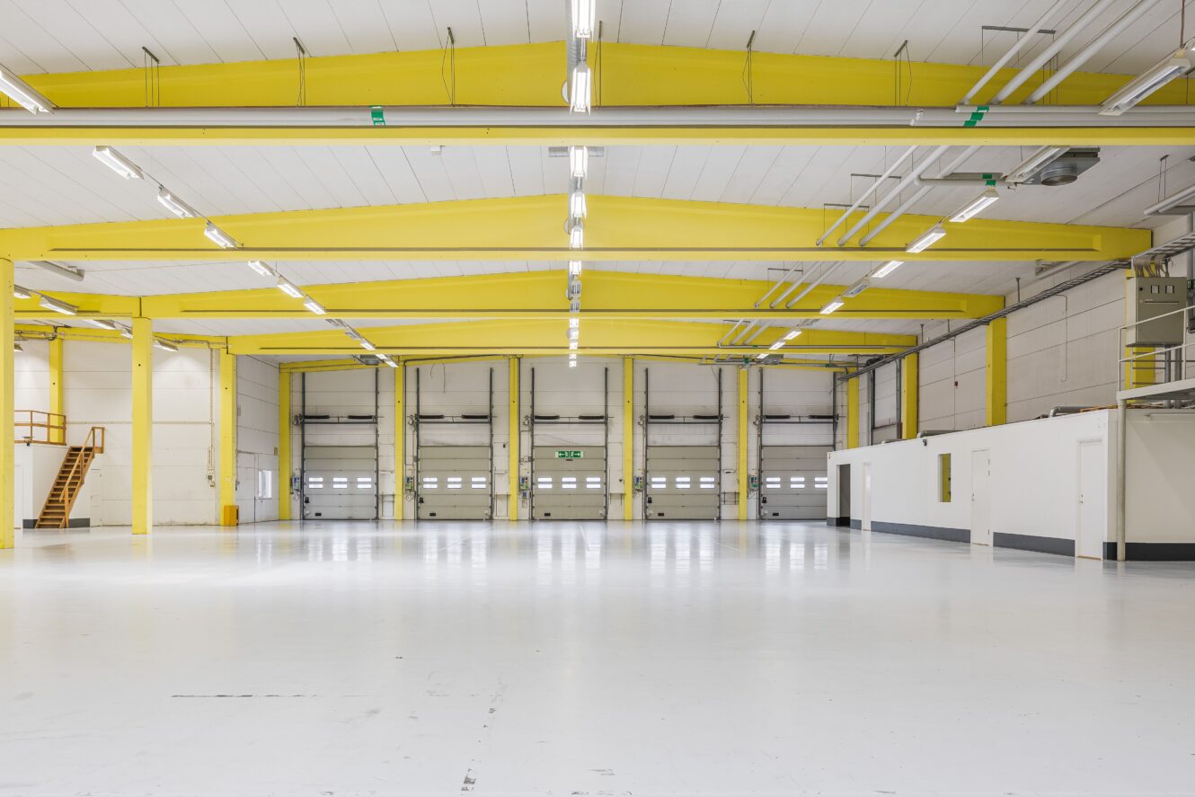 Stor, tom lagerlokal med vita golv, gula takbjälkar, dörrar till lastkajer, lysrör och en trappa på vänster sida.