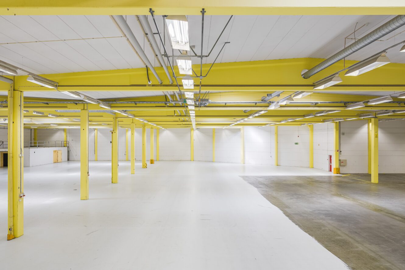 Stor tom lagerlokal med gula bärande balkar, vita väggar och ett golv som delvis är av betong och delvis vitt. Taklampor och exponerade rör syns i taket.