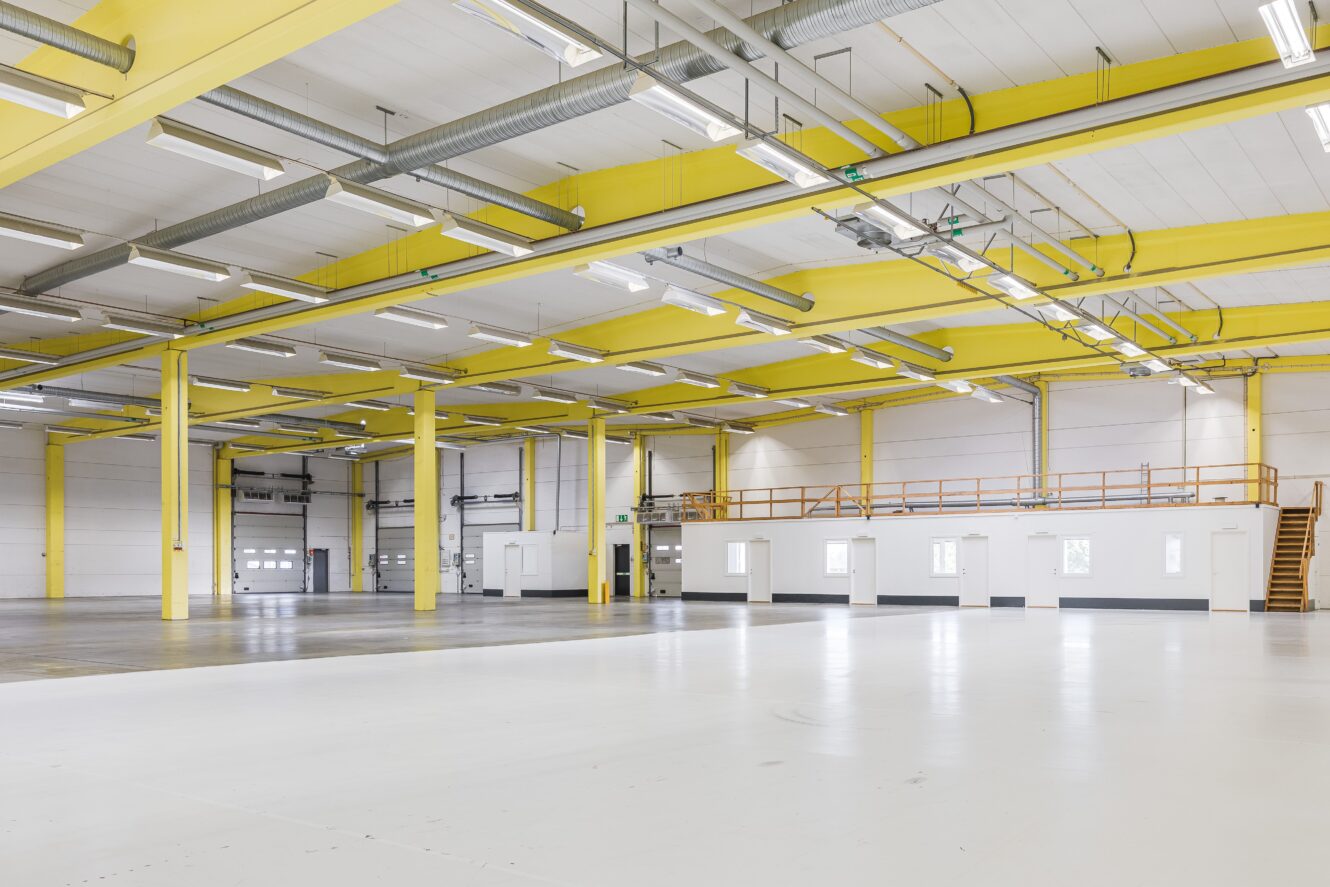 Rymlig, tom industrilokal med gula takbjälkar, vita väggar och en mezzaninvåning med fönster och trappa.